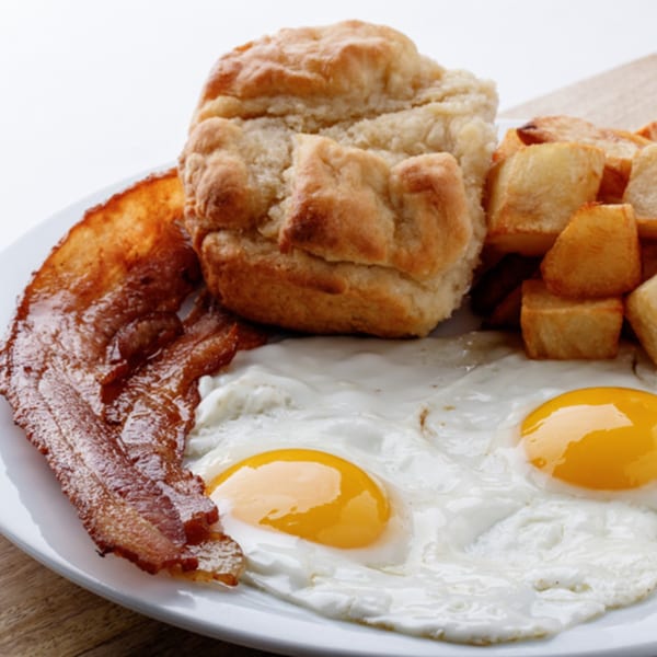 A breakfast plate with crispy bacon, two fried eggs, golden home fries, and a fluffy biscuit.