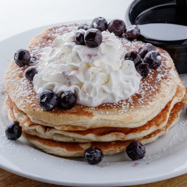 Stack of pancakes topped with whipped cream, blueberries, and powdered sugar, with syrup on the side.